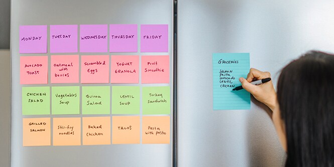 A refrigerator with Post-it® Notes showing a weekly meal plan, and a person writing a grocery list on a lined Post-it® Note

