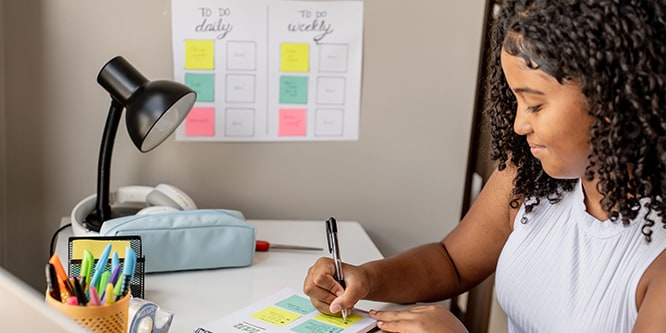 Student making notes on Post-it® Notes, with a revision timetable in the background detailing daily and weekly tasks
