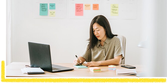 Woman sitting at the desk, making a work to-do list and using Post-it® Notes
