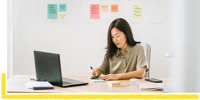 Woman sitting at the desk, making a work to-do list and using Post-it® Notes
